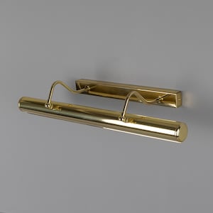 May include: A polished gold-tone metal picture light fixture featuring a long cylindrical lamp head attached to a rectangular wall-mounting plate by two curved, adjustable support arms. The finish is reflective and smooth throughout the entire unit.