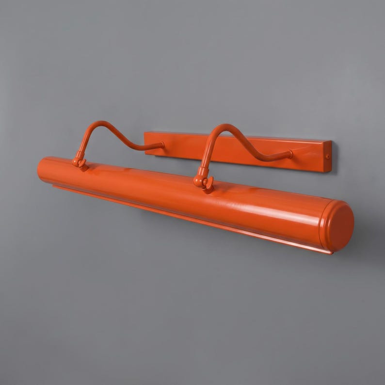 May include: A bright orange picture light with a long cylindrical lamp head and a rectangular wall-mounting plate. The fixture features two curved, adjustable arms that connect the lamp head to the base, allowing for directional light positioning.