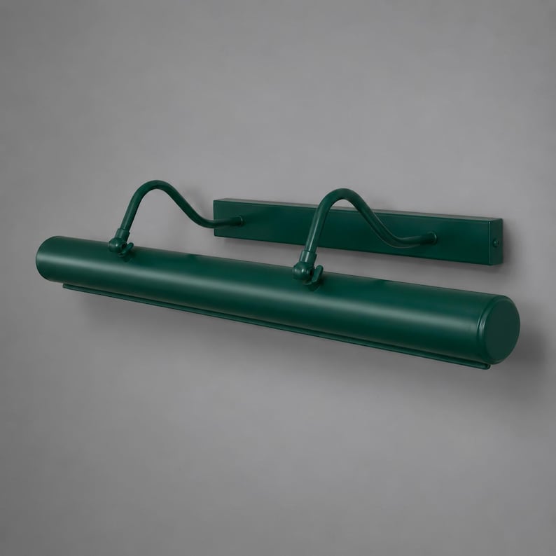 May include: A dark green metal picture light featuring a long cylindrical lamp head attached to a rectangular wall-mounting plate by two curved, adjustable arms. The fixture has a smooth, matte finish and a streamlined, horizontal design.