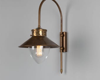 May include: A brass wall sconce in a vintage style. The light fitting has a conical shade, a clear glass globe, and a curved arm. The sconce is mounted on a rectangular backplate and features an exposed light bulb.
