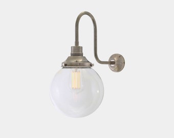 Può includere: Un'applique da parete in stile vintage con un paralume in vetro trasparente a forma di globo. L'apparecchio ha un braccio metallico curvo e una piastra di montaggio circolare. La lampadina è visibile all'interno del vetro.