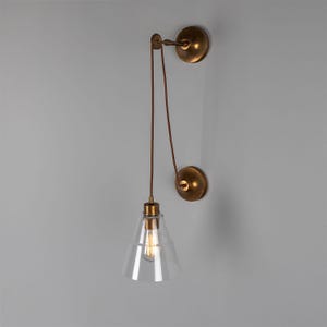 May include: A brass wall-mounted light fixture with a clear glass cone shade and a pull chain. The light fixture is mounted on a white wall.
