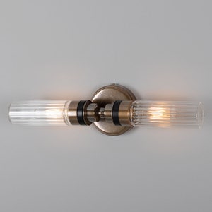 May include: A brushed brass wall sconce with two clear glass cylinder shades. The shades have a ribbed texture and are illuminated by light bulbs.