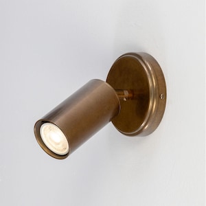 May include: A bronze-colored, wall-mounted spotlight fixture. The cylindrical light head is angled, with a bright light emanating from the opening. The fixture is attached to a circular base.