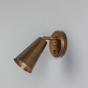 May include: A brass wall-mounted spotlight with a cone-shaped shade and a round base. The light has a switch on the base.