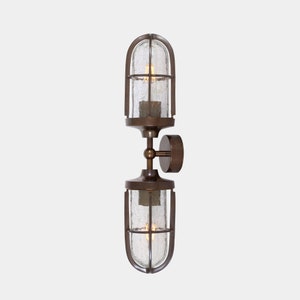 Puede incluir: Aplique de pared doble de bronce con un diseño industrial vintage. Cuenta con dos pantallas de vidrio texturizado encerradas en una jaula de metal, una placa de montaje redonda y un brazo de conexión. Para iluminación interior.