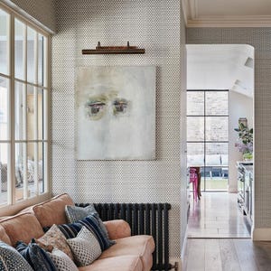 May include: A living space featuring a peach-colored sofa with patterned throw pillows, a black radiator, and a square abstract painting with muted tones hanging on a wall covered in small-scale geometric wallpaper, adjacent to a glass-paned doorway.