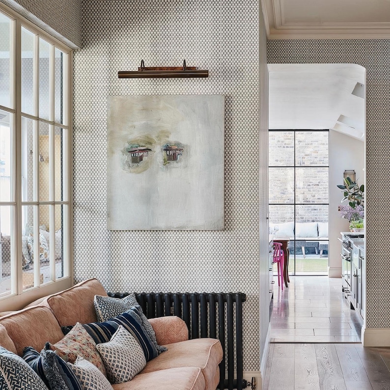 May include: A living space featuring a peach-colored sofa with patterned throw pillows, a black radiator, and a square abstract painting with muted tones hanging on a wall covered in small-scale geometric wallpaper, adjacent to a glass-paned doorway.