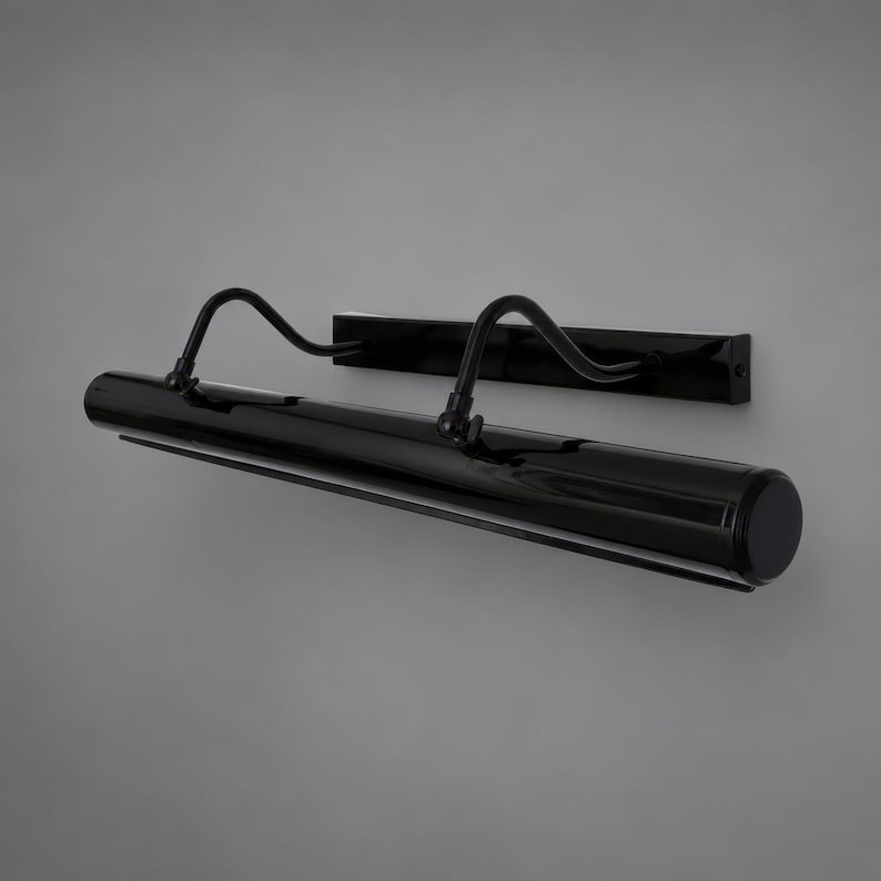 May include: A sleek black picture light featuring a long cylindrical lamp head attached to a rectangular wall-mounting plate by two curved, adjustable metal arms. The entire fixture has a smooth, glossy finish and a minimalist, linear design.