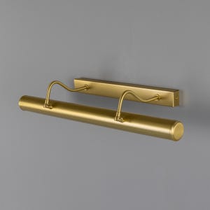 May include: A matte gold picture light fixture featuring a horizontal cylindrical lamp head attached to a rectangular wall-mounting plate by two curved, adjustable metal arms. The design is sleek and functional, providing a focused light source.