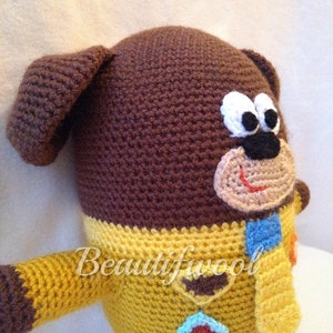 May include: A crocheted dog doll in brown, yellow, and blue. The dog features large white-rimmed eyes, a black nose, and a red smiling mouth. It is wearing a yellow sweater and a blue and orange scarf. The text "Beautifwool" is visible on the doll.