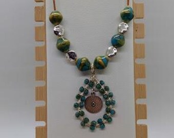 May include: A handcrafted necklace with a brown leather cord, featuring green and silver beads. The pendant is a copper-colored coin surrounded by a teal beaded teardrop shape. The necklace is displayed on a wooden jewelry stand.