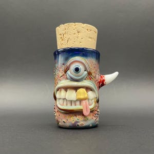 Pode incluir: Uma garrafa de vidro soprado à mão única com uma rolha de cortiça. A garrafa apresenta um design de rosto de monstro caprichoso com um único olho, dentes grandes e uma língua saliente. A paleta de cores inclui detalhes em azul, rosa e dourado.