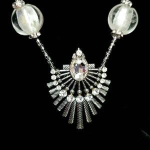 May include: A silver necklace with a large crystal pendant surrounded by smaller crystals. The pendant is shaped like a sunburst with radiating points. The necklace has two large, clear glass beads on either side of the pendant.