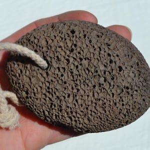Könnte beinhalten: Ein brauner, poröser Bimsstein mit einer cremefarbenen Schnur. Der ovale Stein wird in einer Hand gehalten, wodurch seine strukturierte Oberfläche hervorgehoben wird. Dieses natürliche Fußpflegeprodukt ist zum Peelen bestimmt.