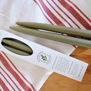 Puede incluir: Una caja de tres velas de taper verdes con el texto "CAPECANDLE BAYBERRY SCENTED TAPERS AS FOLKLORE GOES..." y "SCENTED TAPERS" en la caja. Las velas están sobre una toalla de rayas blancas y rojas.