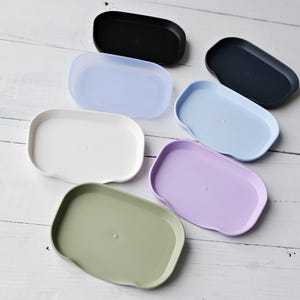 Può includere: Sette vassoi di plastica di diversi colori, in una varietà di forme, su una superficie di legno bianco. I vassoi sono blu chiaro, blu scuro, bianco, verde chiaro, lavanda, nero e trasparente.
