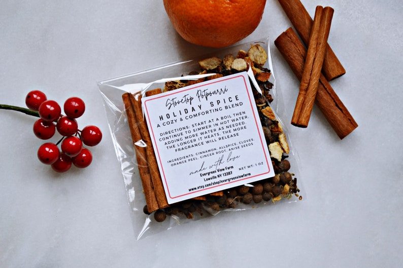 Pu&ograve; includere: Un sacchetto di plastica trasparente pieno di una miscela di spezie per pot-pourri da stufa. L'etichetta recita "Stovetop Potpourri Holiday Spice A Cozy & Comforting Blend" con istruzioni e ingredienti elencati. Il sacchetto &egrave; su una superficie bianca con un rametto di bacche rosse e un'arancia intera a lato.