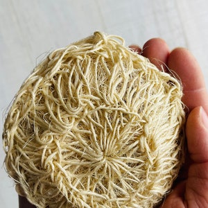 Puede incluir: Primer plano de una esponja de luffa natural, hecha de fibras vegetales secas, en una mano. La luffa es de color beige claro y tiene una apariencia texturizada y tejida.