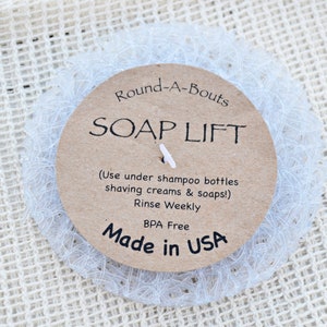 May include: A white mesh soap saver with a brown paper label that reads "Round-A-Bouts SOAP LIFT (Use under shampoo bottles shaving creams & soaps!) Rinse Weekly BPA Free Made in USA"