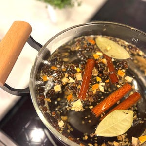 Pu&ograve; includere: Una pentola di vetro con un manico di legno &egrave; posizionata su un piano cottura nero. La pentola &egrave; piena di acqua e varie spezie, tra cui bastoncini di cannella, foglie di alloro e grani di pepe nero.