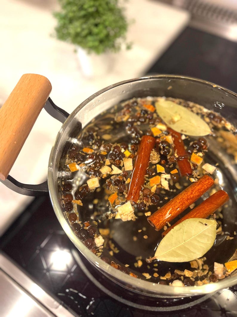 Pu&ograve; includere: Una pentola di vetro con un manico di legno &egrave; posizionata su un piano cottura nero. La pentola &egrave; piena di acqua e varie spezie, tra cui bastoncini di cannella, foglie di alloro e grani di pepe nero.