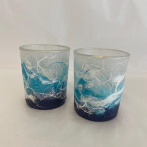 May include: Two decorative glass votive candle holders. The holders feature a design with shades of blue and white, resembling a watercolour painting. The base is a deep navy blue, transitioning to lighter blues and white.
