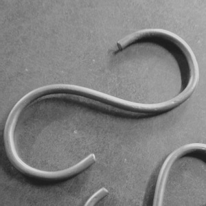 May include: Three silver S-shaped hooks, probably for hanging items, are arranged on a grey surface.