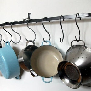 May include: A black metal pot rack with hooks hanging from the ceiling. There are five pots and pans hanging from the hooks, including a blue pot, a silver colander, and a silver pot.
