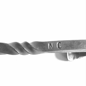 Puede incluir: Primer plano de una varilla de metal retorcida con las letras "NC" estampadas en el extremo.