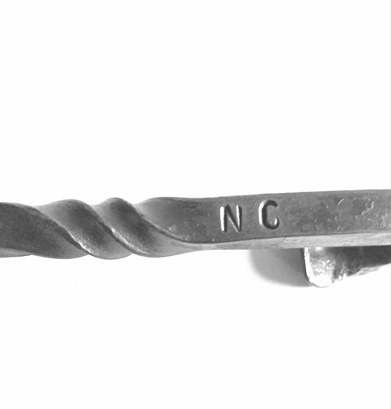 Puede incluir: Primer plano de una varilla de metal retorcida con las letras "NC" estampadas en el extremo.