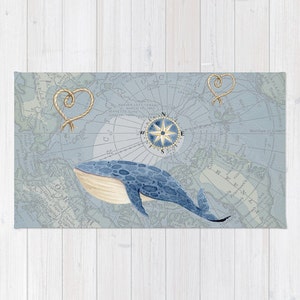May include: A rectangular, nautical-themed rug featuring a blue whale illustration. The rug has a light blue background with a vintage map design, a compass rose, and rope heart accents. The whale is blue with a cream-colored belly.