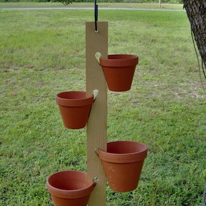 May include: A hanging wooden planter with four terracotta pots. The pots are suspended from a vertical wooden plank with heart-shaped cutouts. The planter hangs from a black cord, suitable for outdoor use in a garden or patio setting.