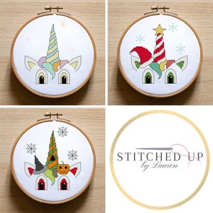 May include: Three cross-stitch embroidery hoops with different unicorn designs. The first hoop features a unicorn with a rainbow horn and a blue and white mane. The second hoop features a unicorn with a Christmas tree hat and a red and green striped horn. The third hoop features a unicorn with a witch hat and a pumpkin horn. The hoops are all on a wooden surface.