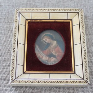 May include: A framed oval painting of a woman holding a baby. The woman is wearing a red robe and a blue head covering. The painting is set against a red velvet background.