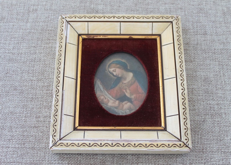 May include: A framed oval painting of a woman holding a baby. The woman is wearing a red robe and a blue head covering. The painting is set against a red velvet background.