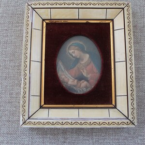 May include: A framed oval painting of a woman holding a baby. The woman is wearing a blue head covering and a red robe. The painting is set against a burgundy velvet background.