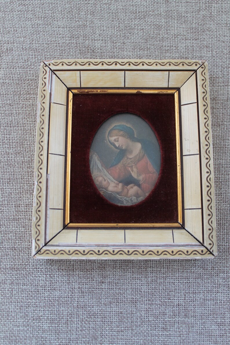May include: A framed oval painting of a woman holding a baby. The woman is wearing a blue head covering and a red robe. The painting is set against a burgundy velvet background.