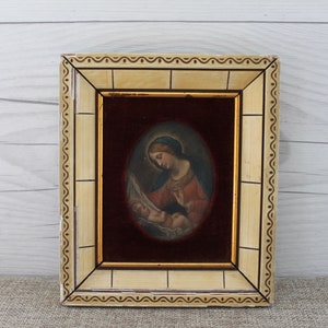 May include: A framed painting of the Virgin Mary holding the baby Jesus. The painting is set against a red velvet background and is framed in a cream-colored frame with gold accents.