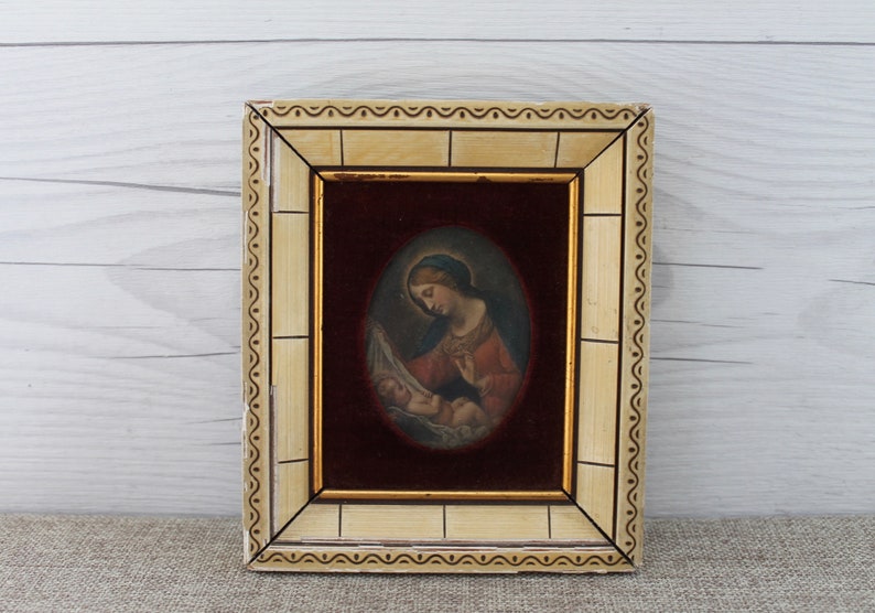 May include: A framed painting of the Virgin Mary holding the baby Jesus. The painting is set against a red velvet background and is framed in a cream-colored frame with gold accents.