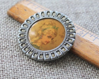 May include: A silver-toned brooch with a white enamel border and a round cameo featuring a portrait of a woman with blonde hair. The cameo is set in a silver-toned frame with a decorative border.