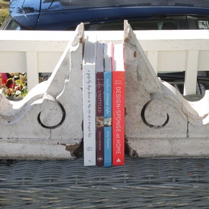 May include: A set of white painted wooden bookends with a decorative scroll design, holding up four books with titles including "Design-Sponge at Home", "Little Unstyled", and "A perfectly divine home is a sign of a magnificent life".