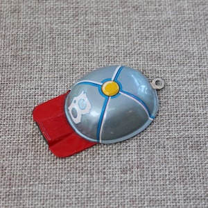 May include: A vintage metal toy helmet with a red visor. The helmet is primarily light blue with white, yellow, and blue accents. A small, white cartoon character is on the side. A small metal loop is attached.