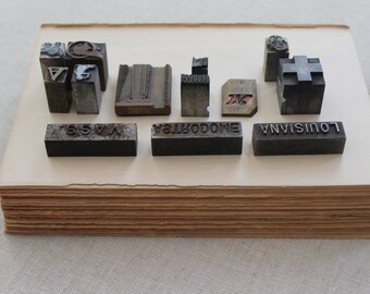 May include: A collection of vintage metal letterpress printing blocks, some with letters and symbols, arranged on a stack of aged book pages. The blocks are various shapes and sizes, with visible wear and patina, and the word "LOUISIANA" is visible.