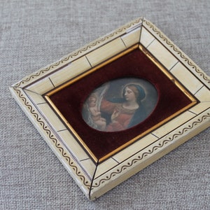 May include: A small oval-shaped framed painting of a woman holding a child. The frame is white with gold accents and a burgundy velvet mat.