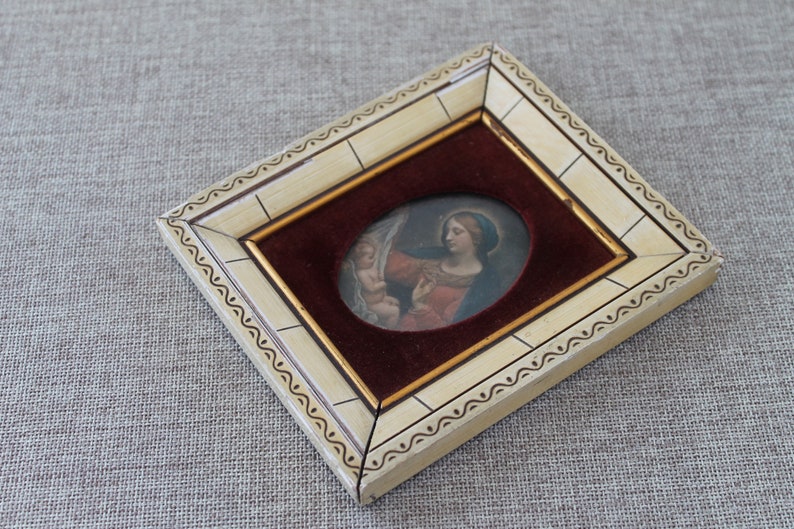 May include: A small oval-shaped framed painting of a woman holding a child. The frame is white with gold accents and a burgundy velvet mat.