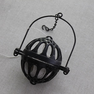 May include: A black metal hanging incense burner with a spherical cage design. The burner has a hinged opening and a chain for hanging. A white string is attached to the burner. The background is a light gray fabric.