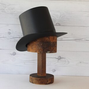 May include: A black top hat with a wide brim sits on a wooden hat form. The hat form is on a wooden base.