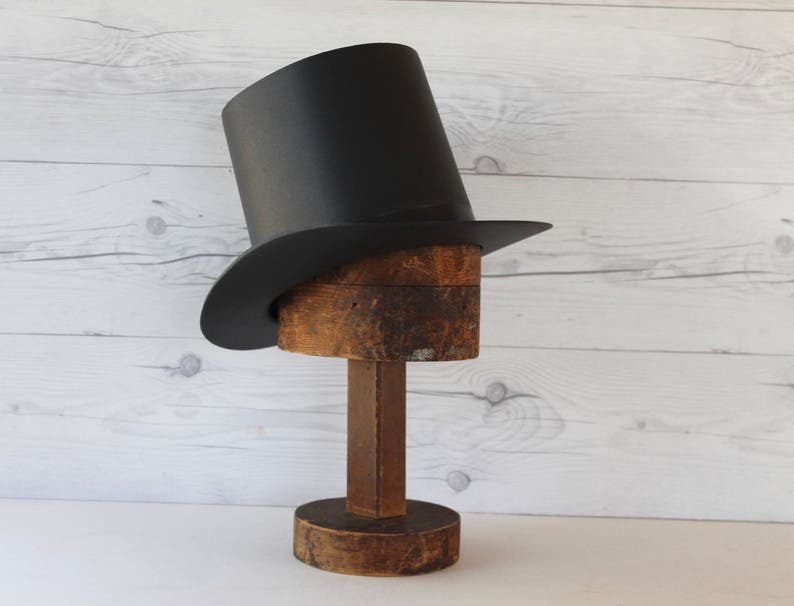 May include: A black top hat with a wide brim sits on a wooden hat form. The hat form is on a wooden base.