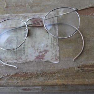 May include: Silver round-framed glasses with a thin metal frame and clear lenses. The glasses are lying on a worn, brown fabric surface.